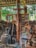 A collection of religious icons, paintings, and a large wooden cross are arranged against a rustic backdrop of wooden beams and stone. The setting appears to be outdoors or in a partially enclosed space, with surrounding greenery visible. The various images depict Christian religious figures and scenes, carefully displayed and propped up.