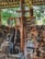 A collection of religious icons, paintings, and a large wooden cross are arranged against a rustic backdrop of wooden beams and stone. The setting appears to be outdoors or in a partially enclosed space, with surrounding greenery visible. The various images depict Christian religious figures and scenes, carefully displayed and propped up.