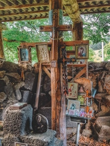 A collection of religious icons, paintings, and a large wooden cross are arranged against a rustic backdrop of wooden beams and stone. The setting appears to be outdoors or in a partially enclosed space, with surrounding greenery visible. The various images depict Christian religious figures and scenes, carefully displayed and propped up.