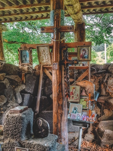 A collection of religious icons, paintings, and a large wooden cross are arranged against a rustic backdrop of wooden beams and stone. The setting appears to be outdoors or in a partially enclosed space, with surrounding greenery visible. The various images depict Christian religious figures and scenes, carefully displayed and propped up.