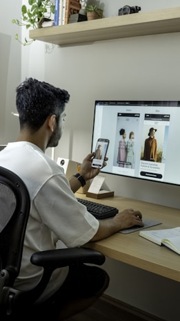 A person is seated at a desk using a computer and a smartphone simultaneously, possibly comparing fashion images on both screens. The computer monitor displays a fashion-related website with large images of models and text about fashion collections. The setting includes a modern desk with office supplies, a notepad, and a potted plant.