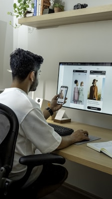 A person is seated at a desk using a computer and a smartphone simultaneously, possibly comparing fashion images on both screens. The computer monitor displays a fashion-related website with large images of models and text about fashion collections. The setting includes a modern desk with office supplies, a notepad, and a potted plant.