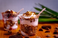 Two dessert cups filled with layers of ice cream, crumbled cookies, and a toasted marshmallow topping. The cups are placed on a wooden surface with green leaves in the background. Chocolate pieces and cookie crumbs are scattered around.
