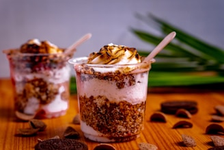 Two dessert cups filled with layers of ice cream, crumbled cookies, and a toasted marshmallow topping. The cups are placed on a wooden surface with green leaves in the background. Chocolate pieces and cookie crumbs are scattered around.