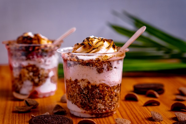 Two dessert cups filled with layers of ice cream, crumbled cookies, and a toasted marshmallow topping. The cups are placed on a wooden surface with green leaves in the background. Chocolate pieces and cookie crumbs are scattered around.