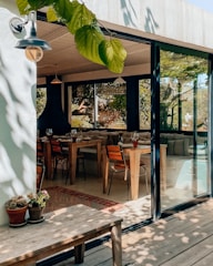 A cozy indoor dining area with wooden furniture is visible through large glass doors. Several tables are set with glasses and cutlery, and the space is well-lit with natural light filtering through the leaves outside. Potted plants add a touch of greenery to the scene on a wooden bench in the foreground.