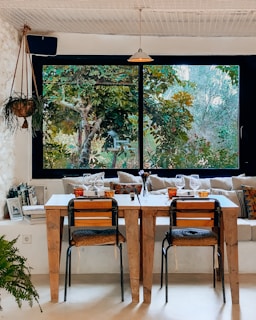 A cozy indoor dining area with two wooden tables and chairs, set against a large window that offers a view of lush green foliage. The tables are set with wine glasses and small centerpieces, and the space is decorated with a hanging plant and colorful cushions on a bench. Books are stacked neatly against the wall, adding a homely touch.