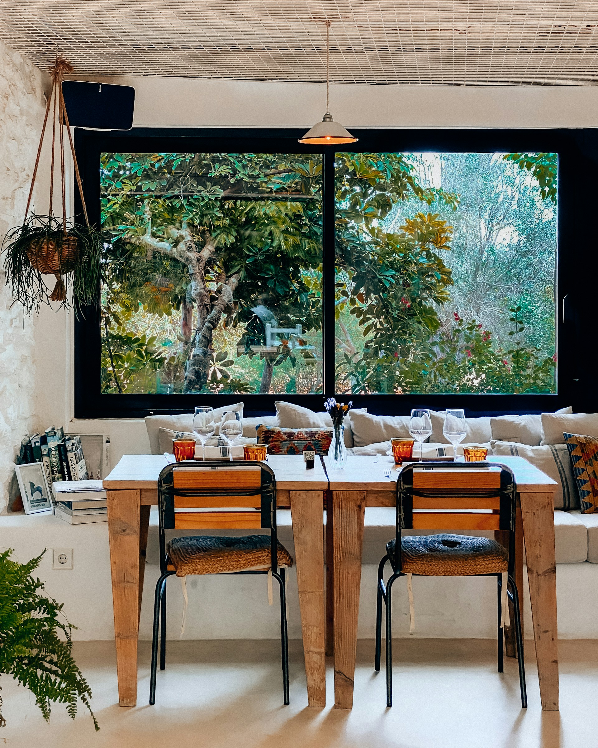 A cozy indoor dining area with two wooden tables and chairs, set against a large window that offers a view of lush green foliage. The tables are set with wine glasses and small centerpieces, and the space is decorated with a hanging plant and colorful cushions on a bench. Books are stacked neatly against the wall, adding a homely touch.