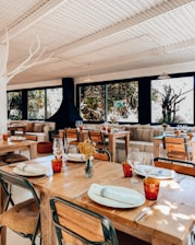 A cozy restaurant interior with wooden tables set with cutlery and plates, adorned with red glasses. The seating includes metal and wooden chairs and comfortable couches with colorful cushions. Large windows provide a view of lush greenery outside, allowing natural light to fill the room. The atmosphere is enhanced by decorative elements like a vase of dried flowers on the table and a modern, black fireplace.