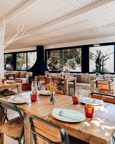 A cozy restaurant interior with wooden tables set with cutlery and plates, adorned with red glasses. The seating includes metal and wooden chairs and comfortable couches with colorful cushions. Large windows provide a view of lush greenery outside, allowing natural light to fill the room. The atmosphere is enhanced by decorative elements like a vase of dried flowers on the table and a modern, black fireplace.