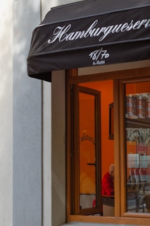 A storefront with a black awning displaying the word 'Hamburguesería'. The window reveals an interior painted in warm tones, possibly a cafe, with a person wearing a red top seated inside.