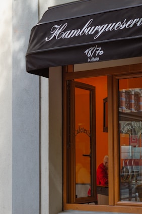 A storefront with a black awning displaying the word 'Hamburguesería'. The window reveals an interior painted in warm tones, possibly a cafe, with a person wearing a red top seated inside.