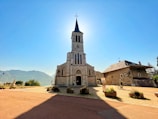 There is a stone church with a tall steeple illuminated by sunlight from behind, casting a shadow on the foreground. To the right, an adjacent building with a sloped roof and windows can be seen. The scene is surrounded by a wide open area with planters and flags to the left, with mountains visible in the distant background. A cyclist is present near the building.
