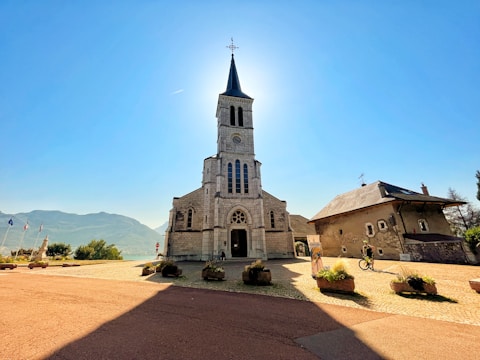 There is a stone church with a tall steeple illuminated by sunlight from behind, casting a shadow on the foreground. To the right, an adjacent building with a sloped roof and windows can be seen. The scene is surrounded by a wide open area with planters and flags to the left, with mountains visible in the distant background. A cyclist is present near the building.
