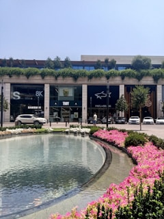 An outdoor landscaped area with a large curved water feature surrounded by pink and white flowers. In the background is a modern building with storefronts, including visible signs for Samsung, Troy, and Paul & Shark. Several cars are parked nearby, with trees and greenery decorating the rooftop of the building.