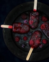 Four dark red popsicles are arranged on a round, black plate, surrounded by scattered blackberries and blueberries. The popsicles have a rich, berry-infused appearance, with visible fruit pieces embedded inside. The plate is set against a deep, dark background, enhancing the vibrant colors of the berries and popsicles.