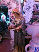 A statue of an angel adorned with luxury brand logos, including Louis Vuitton. The angel holds a pair of vintage telephones and rosary beads, with large wings featuring the Chanel logo. The background is filled with eclectic and colorful neon lights and a variety of objects.