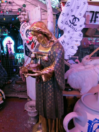 A statue of an angel adorned with luxury brand logos, including Louis Vuitton. The angel holds a pair of vintage telephones and rosary beads, with large wings featuring the Chanel logo. The background is filled with eclectic and colorful neon lights and a variety of objects.