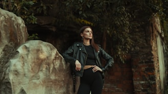 A woman stands confidently with one hand on her hip and the other resting on a large rock. She is wearing a black leather jacket and black clothing, set against a background of stone and foliage. The scene has a moody and somewhat mysterious atmosphere.