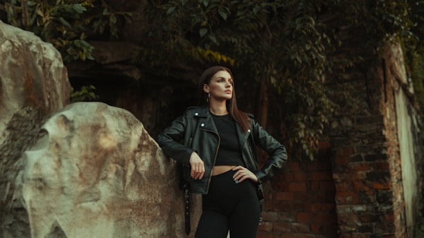 A woman stands confidently with one hand on her hip and the other resting on a large rock. She is wearing a black leather jacket and black clothing, set against a background of stone and foliage. The scene has a moody and somewhat mysterious atmosphere.