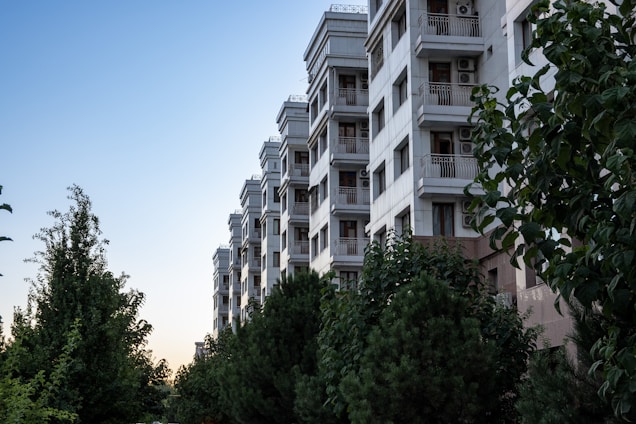 A multi-story residential building with a modern design stands beside a green area filled with various trees and shrubs. The building features balconies with railings and air conditioning units, contrasting against a clear sky as evening approaches.