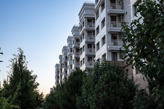 A multi-story residential building with a modern design stands beside a green area filled with various trees and shrubs. The building features balconies with railings and air conditioning units, contrasting against a clear sky as evening approaches.