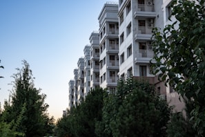 A multi-story residential building with a modern design stands beside a green area filled with various trees and shrubs. The building features balconies with railings and air conditioning units, contrasting against a clear sky as evening approaches.