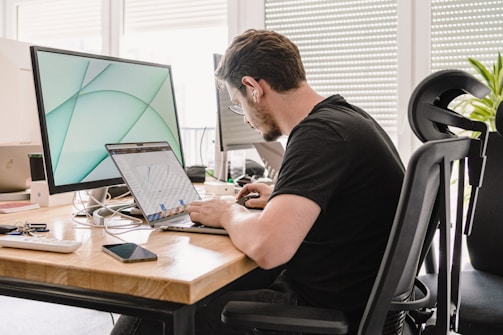 A man is working at a desk equipped with multiple screens, a laptop, and an assortment of office items. He is wearing black clothing, earphones, and glasses while focusing on the laptop in front of him. The work space is modern and bright, with natural light coming from the window blinds behind him.