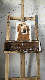 A wooden easel displays a painting of a dog with a brown and white fur. The background is a splattered paint wall with various colors. The easel is equipped with a tray holding various paint smudges.