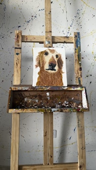 A wooden easel displays a painting of a dog with a brown and white fur. The background is a splattered paint wall with various colors. The easel is equipped with a tray holding various paint smudges.