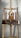 A wooden easel displays a painting of a dog with a brown and white fur. The background is a splattered paint wall with various colors. The easel is equipped with a tray holding various paint smudges.