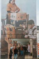 A large mural painting on the side of a building depicts two figures engaged in packing or organizing items in cardboard boxes. The artwork portrays a woman wearing a traditional orange shawl and a necklace, focused on her task, while a boy appears engrossed in his own activity involving a small object. The mural extends above an urban walkway, where people are walking or standing, some dressed warmly, suggesting a chilly environment.