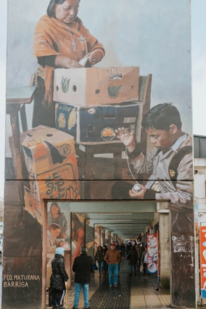 A large mural painting on the side of a building depicts two figures engaged in packing or organizing items in cardboard boxes. The artwork portrays a woman wearing a traditional orange shawl and a necklace, focused on her task, while a boy appears engrossed in his own activity involving a small object. The mural extends above an urban walkway, where people are walking or standing, some dressed warmly, suggesting a chilly environment.
