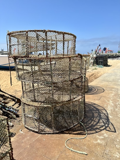 Stacked fishing cages sit on a sandy beach with boats in the background. The area is bright and sunny, featuring clear blue skies and a pathway made of tiles. Flags can be seen in the distance, adding a nautical theme.