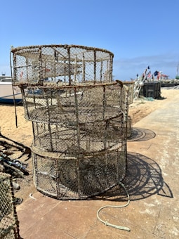 Stacked fishing cages sit on a sandy beach with boats in the background. The area is bright and sunny, featuring clear blue skies and a pathway made of tiles. Flags can be seen in the distance, adding a nautical theme.