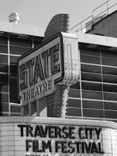 A black and white photograph of a vintage theatre marquee with 'STATE THEATRE' displayed prominently. Below, the words 'TRAVERSE CITY FILM FESTIVAL' are in large letters. The building features classic marquee lighting and signs.