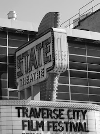 A black and white photograph of a vintage theatre marquee with 'STATE THEATRE' displayed prominently. Below, the words 'TRAVERSE CITY FILM FESTIVAL' are in large letters. The building features classic marquee lighting and signs.