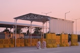 A toll booth or checkpoint structure with a large metal roof and several yellow barriers lined up in front. The setting seems to be outdoors during sunset, with a pink sky and street lamps visible in the background. Trees and a small building or shed can be seen behind the barriers.