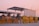 A toll booth or checkpoint structure with a large metal roof and several yellow barriers lined up in front. The setting seems to be outdoors during sunset, with a pink sky and street lamps visible in the background. Trees and a small building or shed can be seen behind the barriers.
