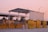 A toll booth or checkpoint structure with a large metal roof and several yellow barriers lined up in front. The setting seems to be outdoors during sunset, with a pink sky and street lamps visible in the background. Trees and a small building or shed can be seen behind the barriers.