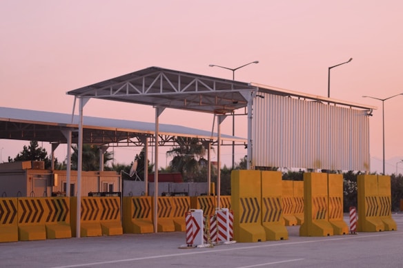 A toll booth or checkpoint structure with a large metal roof and several yellow barriers lined up in front. The setting seems to be outdoors during sunset, with a pink sky and street lamps visible in the background. Trees and a small building or shed can be seen behind the barriers.