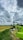 A vibrant green agricultural machine with a cutting mechanism is harvesting crops in a vast golden field under a dramatic sky filled with white and gray clouds. The landscape stretches into the distance with green grass lining the path.