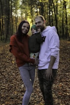 A family of three stands together in a forest during autumn. The ground is covered with fallen leaves, and sunlight filters through the trees, casting a warm glow. The mother wears a red sweater and jeans, while the father is in a white hoodie and camouflage pants. The child, dressed in a green outfit, is being held by the mother.
