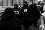 Several people walk together on a busy urban street, with two prominently dressed in NYPD Counterterrorism jackets in the center. The background features tall city buildings.