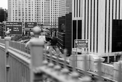 A black and white image depicting an urban scene in Manhattan. Several road signs are visible, including directions to Park Row, FDR Drive, and a welcoming sign for Manhattan. The foreground includes a blurred fence, while the background shows tall buildings with numerous windows.