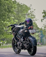A motorcycle with sleek design and a modern helmet placed on the rear seat is parked on a road. The background is lush with greenery, suggesting a serene environment. The motorcycle's registration plate is visible.