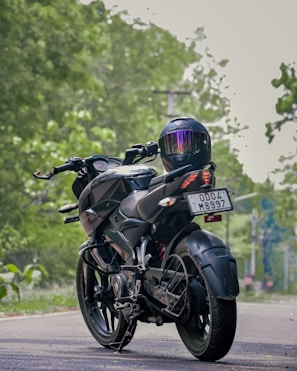 A motorcycle with sleek design and a modern helmet placed on the rear seat is parked on a road. The background is lush with greenery, suggesting a serene environment. The motorcycle's registration plate is visible.
