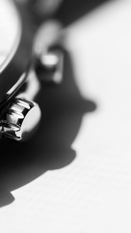 A close-up black and white photograph of a watch crown and pushers, highlighting the intricate metal details. The focus is sharp on the crown with a soft blurred shadow falling on a light surface in the background.