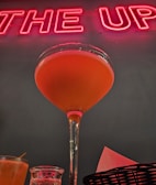 A cocktail in a stemmed glass filled with orange liquid is prominently displayed against a backdrop of red neon lettering. The image includes additional glasses and a basket with napkins at the lower margin, creating a festive and vibrant atmosphere.