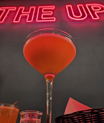A cocktail in a stemmed glass filled with orange liquid is prominently displayed against a backdrop of red neon lettering. The image includes additional glasses and a basket with napkins at the lower margin, creating a festive and vibrant atmosphere.